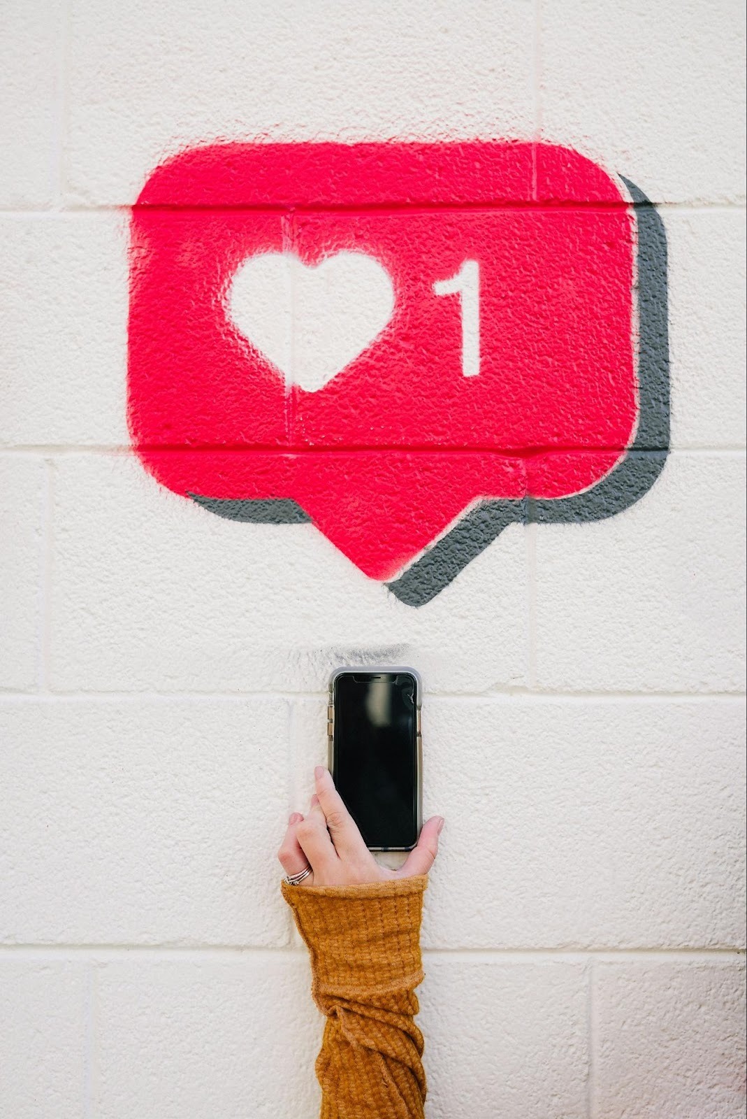 Hand holding a phone near a wall painted with a social media "Like" notification, illustrating audience engagement.